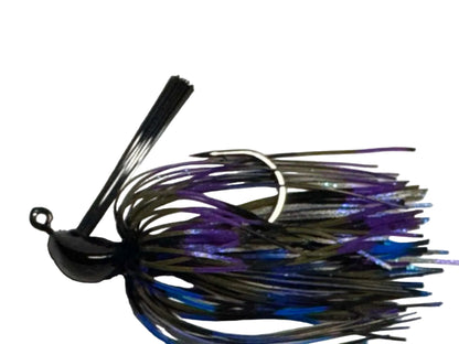 A close-up of a fishing jig lure with a black top, dark metallic skirt, and purple and blue glitter strands, photographed against a plain off-white background.
